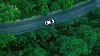 Aerial view of a white car driving along a winding road through dense green forest.