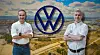 Two Volkswagen employees stand with folded arms in front of a large VW logo over an aerial view of an industrial plant.