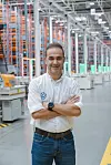 Man in Volkswagen shirt stands with folded arms in a large automated warehouse.
