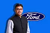 Man in formal attire standing in front of a large Ford logo on a blue background.