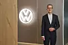 Volkswagen executive in a dark suit stands indoors beside an illuminated VW logo on the wall.