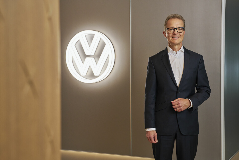 Volkswagen executive in a dark suit stands indoors beside an illuminated VW logo on the wall.