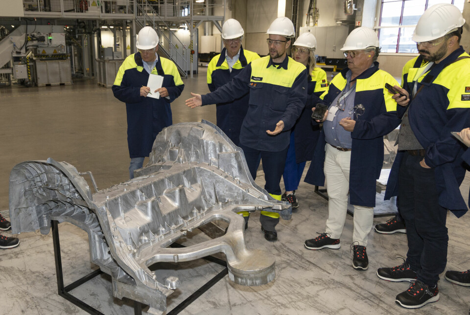 Group in safety gear examining a large aluminium car floor casting on a stand inside a factory.