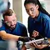 Two workers wearing safety glasses examine a tablet together in a workshop or lab.