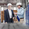 Two men in hard hats talking in a factory aisle beside blue industrial cabinets.