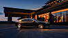 A black luxury sedan parked beside a modern building with warm interior lighting at dusk.