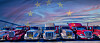A line of coloured heavy-duty trucks parked side by side under a twilight sky with EU stars above.