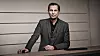 A man in a dark suit stands behind a table against a plain wall.