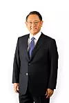 A man in a dark business suit and tie stands against a plain white background.