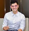 A smiling man wearing pink glasses sits indoors holding a tablet.