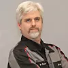 A man with grey hair and a goatee wearing a black work shirt stands against a neutral background.
