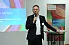 A man in a suit speaks into a microphone beside a presentation screen and a symposium banner.