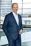 A smiling man in a blue suit stands inside a bright glass-walled showroom with a BMW vehicle visible behind him.