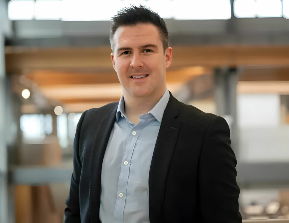 A man in a dark suit stands indoors in front of a softly blurred modern interior with wood beams.
