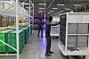 A humanoid robot stands beside stacked crates inside a factory aisle with workers in the background.
