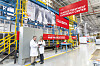 Two people in white coats stand beside a large industrial pretreatment line with red Henkel signs and yellow railings.
