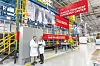 Two people in white coats stand beside a large industrial pretreatment line with red Henkel signs and yellow railings.
