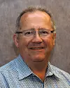 Head-and-shoulders portrait of a man wearing glasses and a blue checked shirt against a grey background.