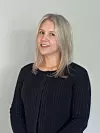 A blonde woman in a black top poses for a portrait against a plain light grey background.