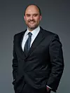 A man in a dark suit and tie stands against a plain grey studio backdrop.