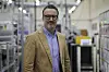 A man in glasses and a tan blazer stands in a factory aisle with machinery and industrial equipment behind him.