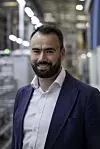 A smiling man in a suit stands in a factory aisle with blurred machinery and bright lights behind him.