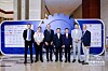 A group of seven people stand on a stage beside a large blue gear-shaped display panel.