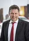 A man in a dark suit and red tie smiles in front of a blurred car and showroom background.