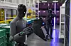 A humanoid robot standing in an industrial facility holds a black component beside green bins and a conveyor line.