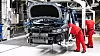 Two workers in red overalls work on a car body on an assembly line inside a factory.
