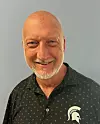 A smiling bald man in a black patterned shirt stands against a plain grey background.