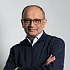 A man wearing glasses and a dark jacket stands with folded arms against a grey backdrop.