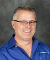 A smiling man in glasses wearing a blue Spraying Systems Co. shirt against a grey background.