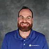 A smiling bearded man in a blue polo shirt stands against a grey studio background.