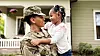 A person in military uniform hugs a smiling child in front of a house.