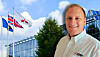 A smiling man in a white shirt stands outdoors in front of flags and a modern office building.