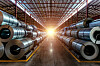 Stacks of shiny steel coils stored on both sides of a warehouse aisle with bright light at the far end.