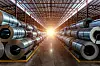 Stacks of shiny steel coils stored on both sides of a warehouse aisle with bright light at the far end.