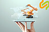 A hand holding a tablet displaying orange industrial robots, charts and network graphics on a pale background.