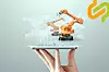 A hand holding a tablet displaying orange industrial robots, charts and network graphics on a pale background.