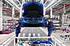 Zwei Beschäftigte stehen vor einem blauen Auto in einer industriellen Montagehalle mit geöffnetem Kofferraum.