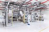 An industrial production line with metal machinery, overhead pipes and a glossy white floor in a bright clean facility.