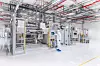An industrial production line with metal machinery, overhead pipes and a glossy white floor in a bright clean facility.