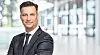 Man in a dark suit stands with arms crossed in a bright modern office interior.
