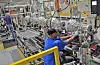 Workers stand beside an automotive assembly line with metal frames, machinery and safety barriers in a factory.