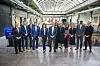 A group of suited business and government leaders stands in a large indoor hall with a curved glass roof.