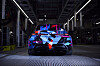 Rear view of a BMW concept car with blue and red geometric livery in a dimly lit factory hall.