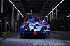 Rear view of a BMW concept car with blue and red geometric livery in a dimly lit factory hall.