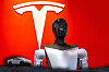 A humanoid robot torso stands in front of a large white Tesla logo on a red background.