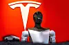 A humanoid robot torso stands in front of a large white Tesla logo on a red background.